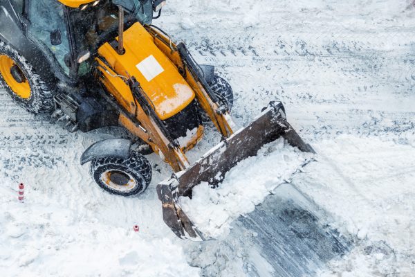 Tractor loader machine uploading dirty snow into dump truck. Cleaning city street, removing snow and ice after heavy snowfalls and blizzard. Snowplow outdoors clean pavement sidewalk road driveway.